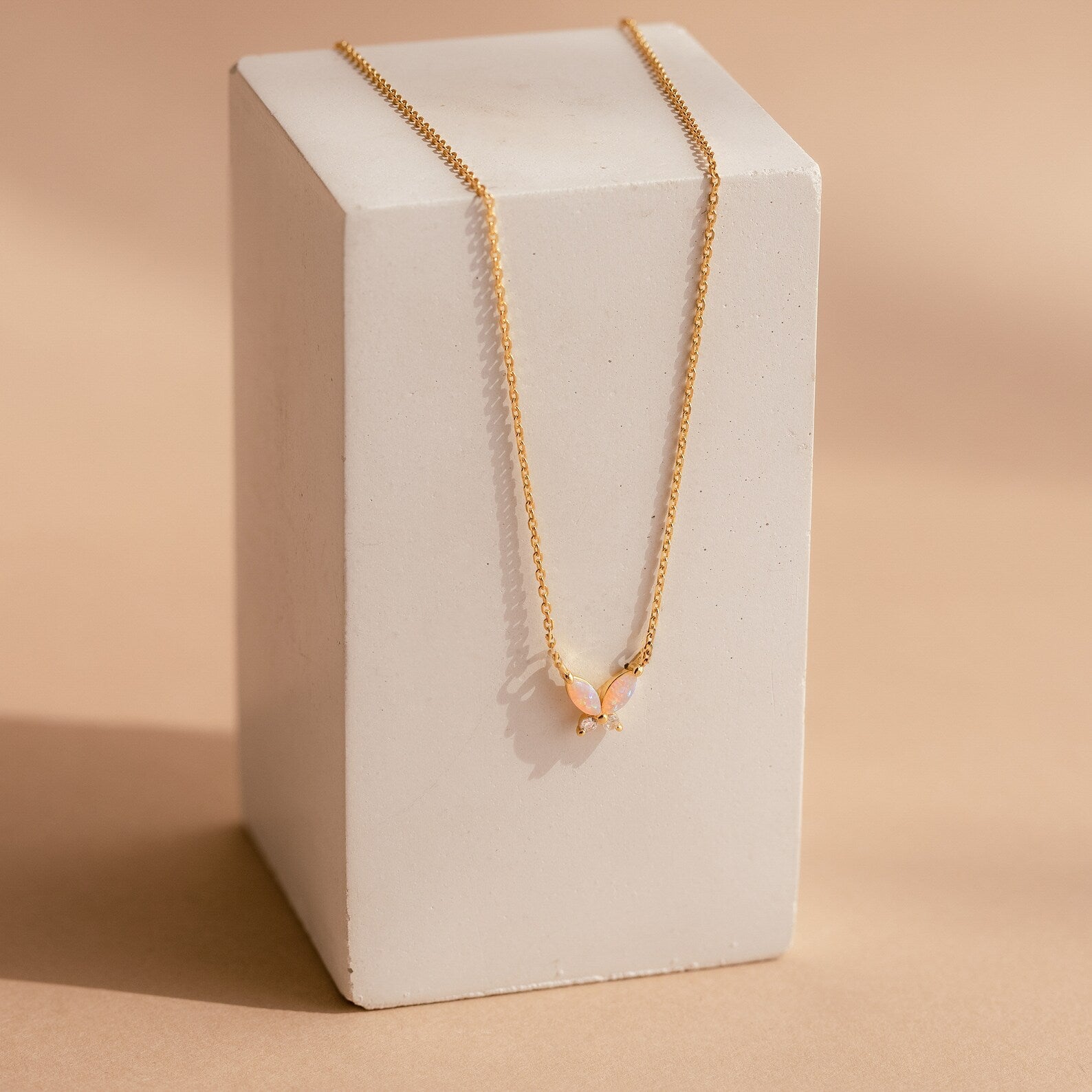 The Pink Opal Butterfly Necklace features a gold chain with a delicate pink opal butterfly pendant, beautifully displayed on a rectangular white block for an elegant touch.