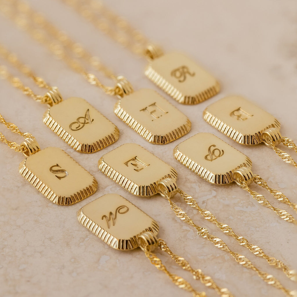 The Traditional Ribbed Initial Necklace features ornate engraved initials on a rectangular charm, elegantly displayed on a light surface.