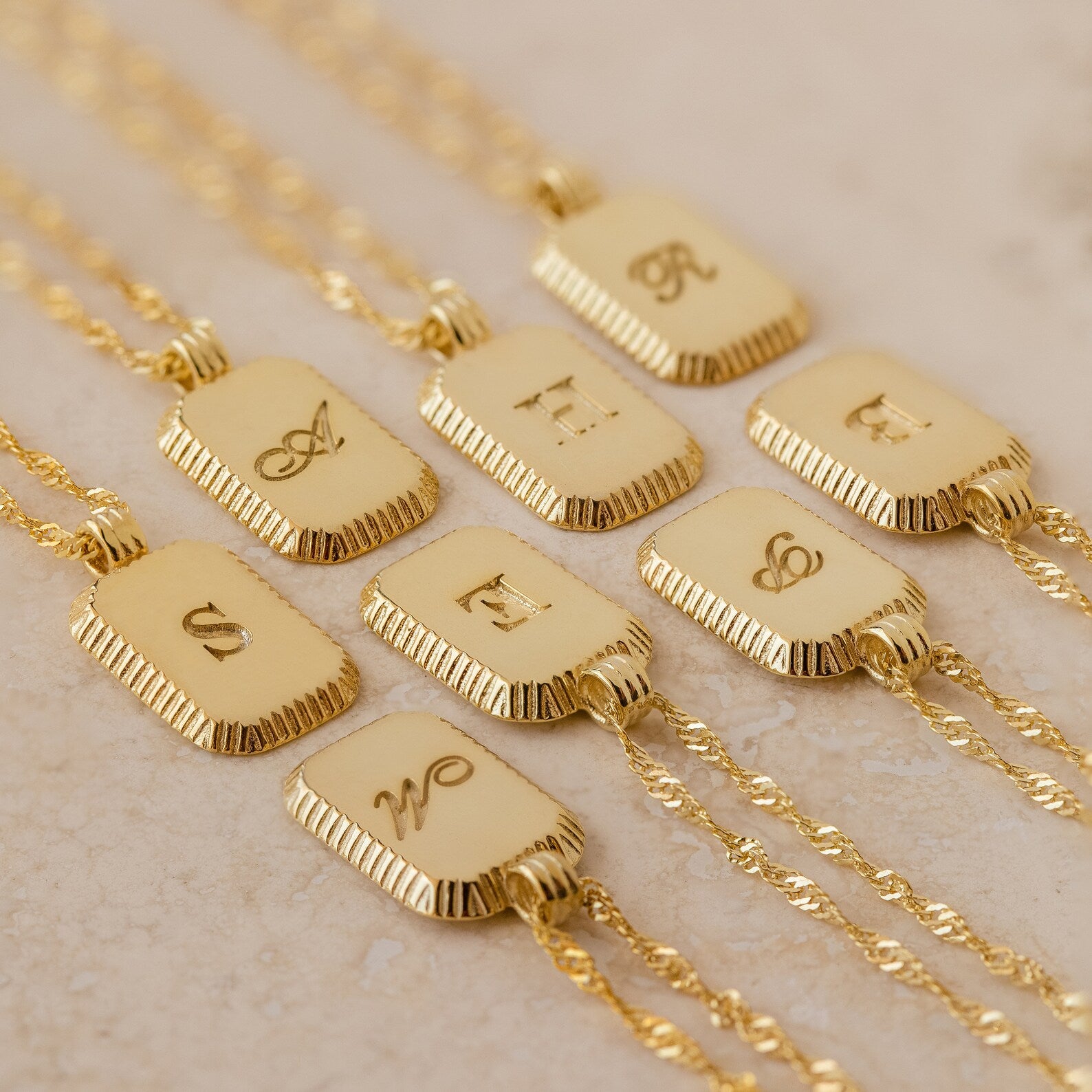 The Traditional Ribbed Initial Necklace features ornate engraved initials on a rectangular charm, elegantly displayed on a light surface.