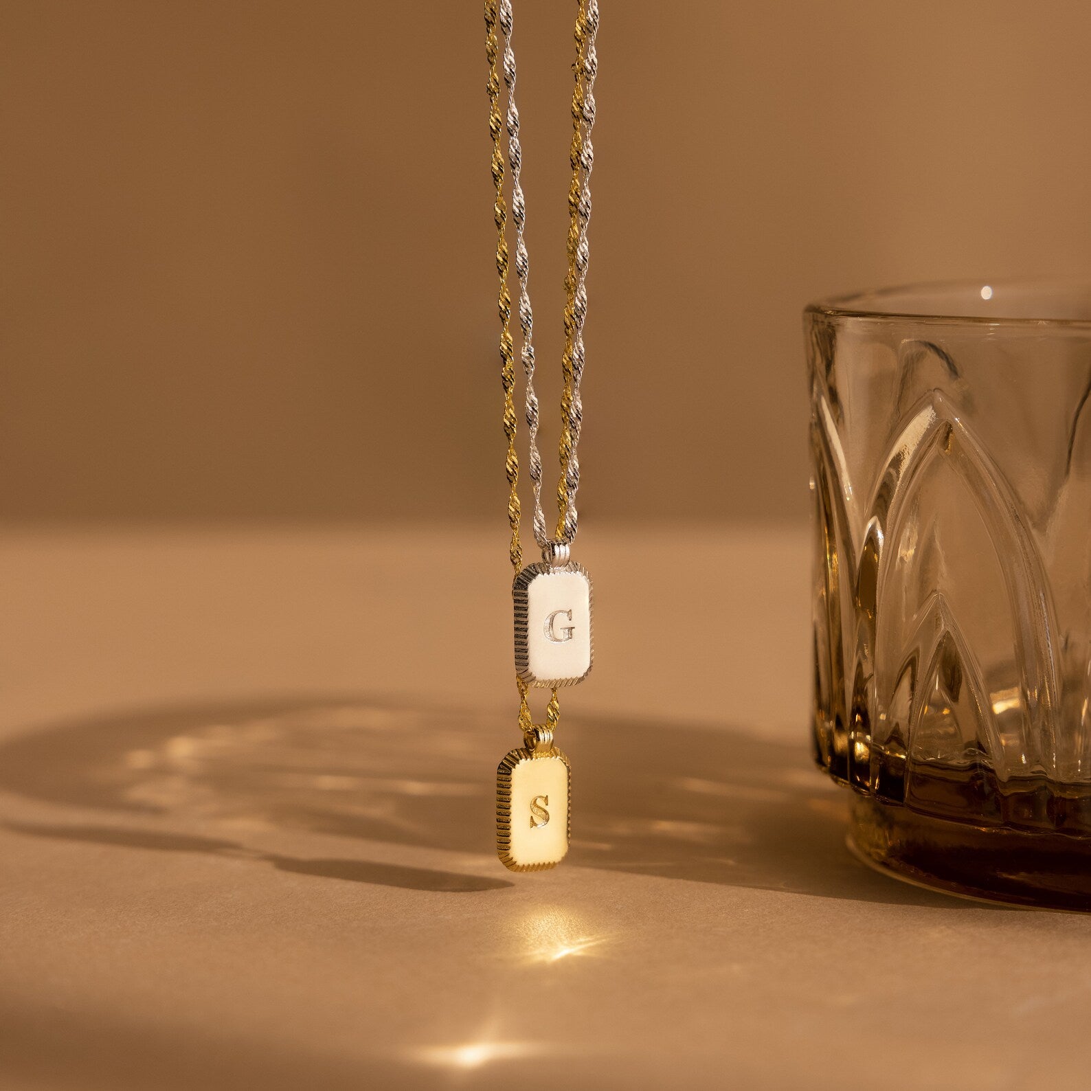 Two Newsletter Ribbed Initial Necklaces in gold and silver engraved with initials hang next to a textured glass on a beige surface under warm lighting.