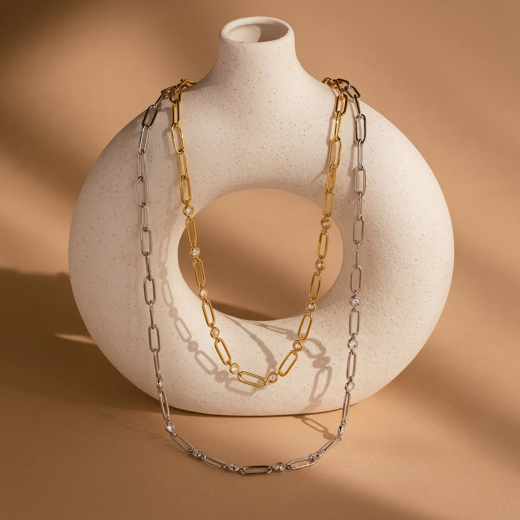 The Diamond Paperclip Necklace, a gold chain featuring minimalist and statement design, drapes over a round white ceramic vase on a beige background.