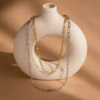The Diamond Paperclip Necklace, a gold chain featuring minimalist and statement design, drapes over a round white ceramic vase on a beige background.