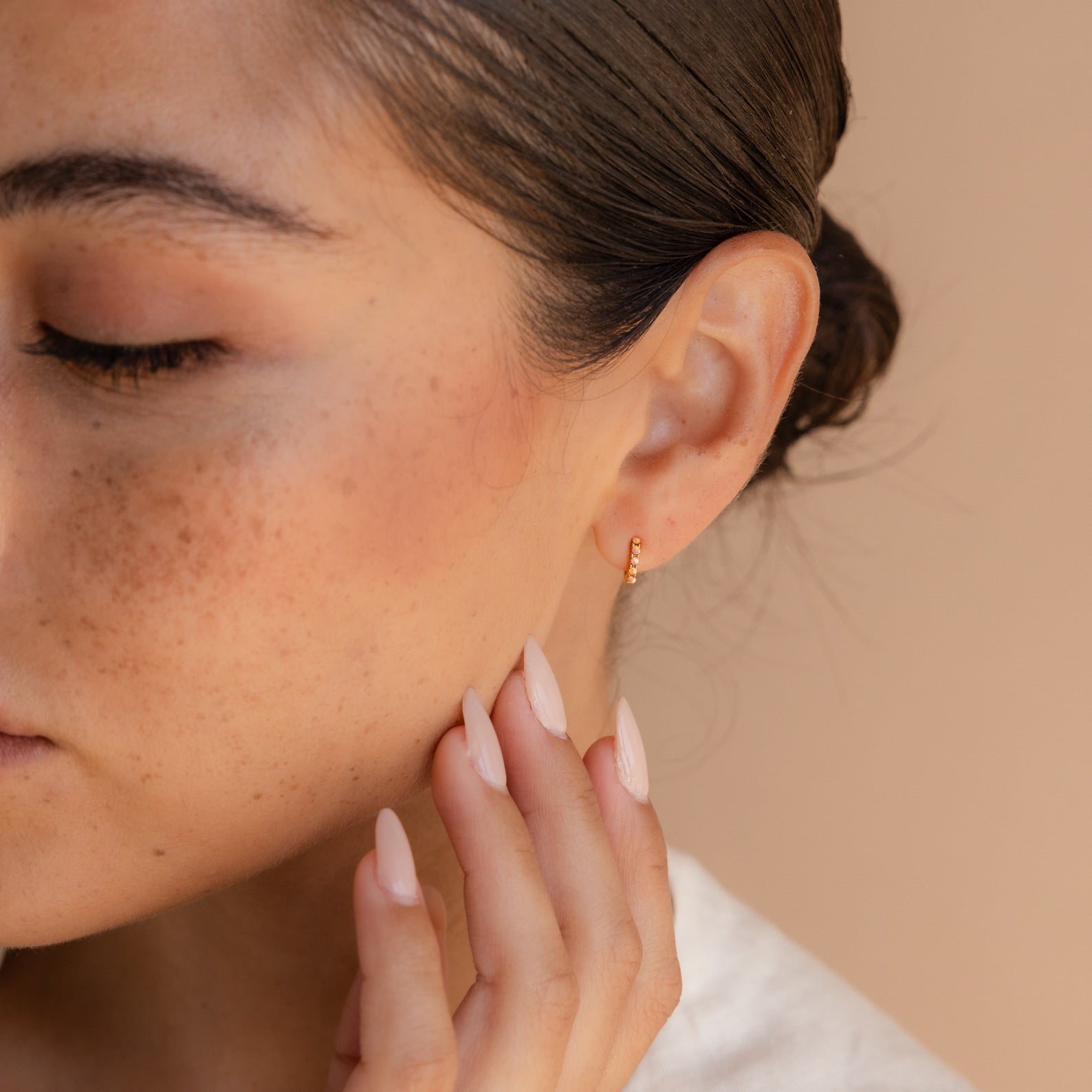 A woman with freckles touches her face, showcasing dainty Pink Opal Huggies and nude manicured nails—a subtle nod to feminine jewelry.