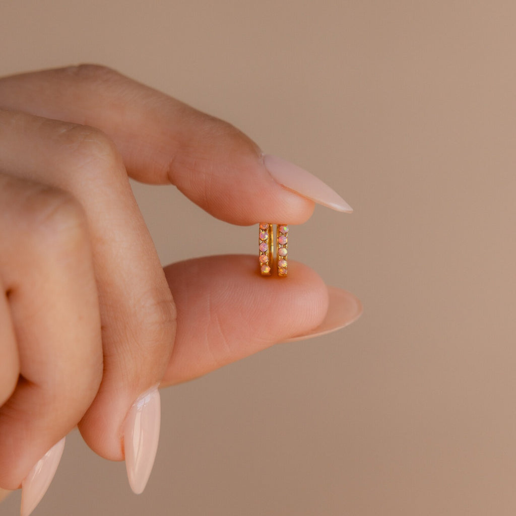 A manicured hand holds a Pink Opal Huggies earring, highlighting its delicate design and shimmering gemstones—an elegant piece of feminine jewelry.
