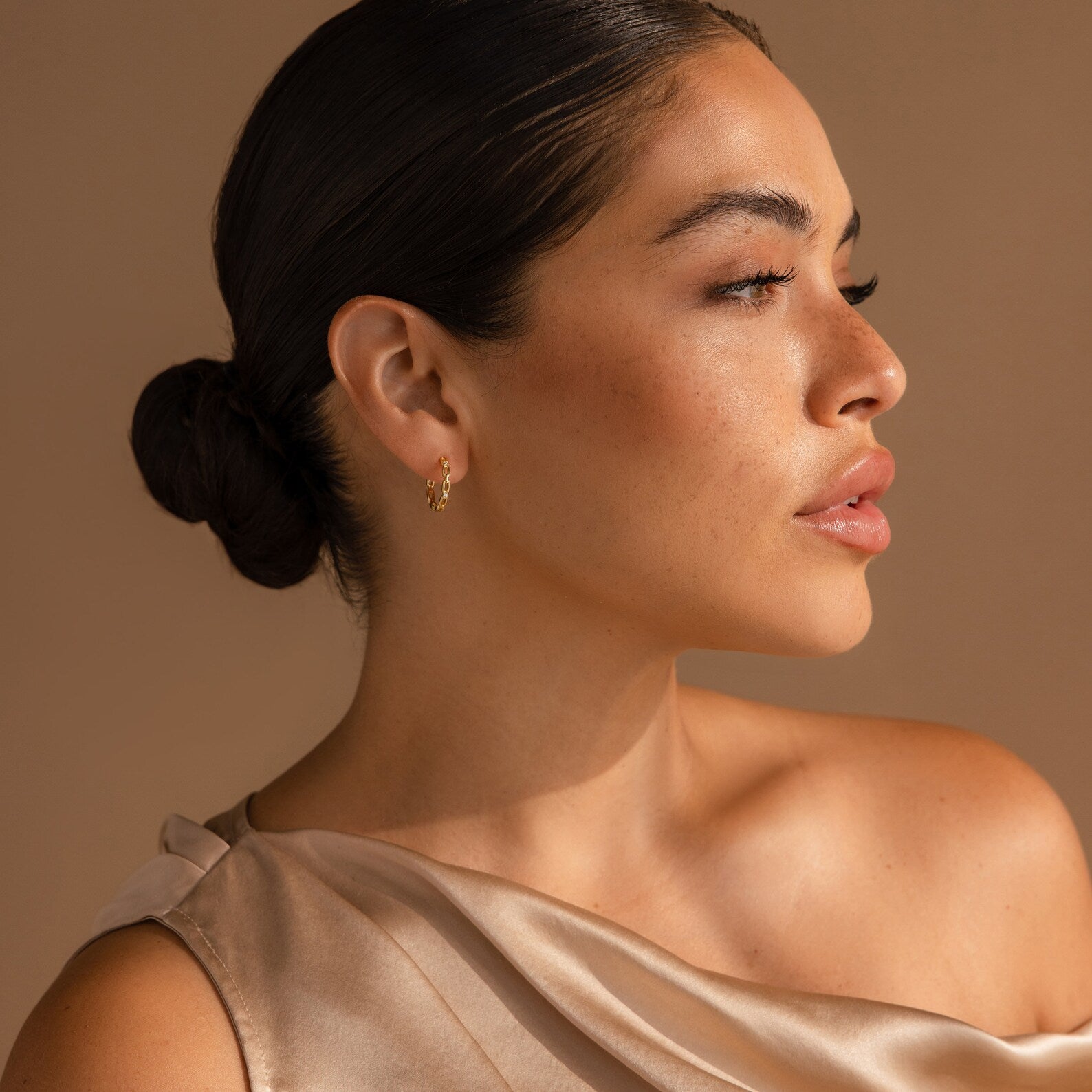 Woman with dark hair in a low bun, wearing Diamond Link Hoops and a satin off-shoulder top, looking to the right.