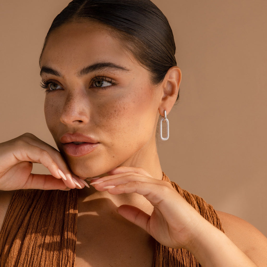 A woman with slicked-back hair poses with her hands near her face, wearing a brown top and Pave Link Hoops.