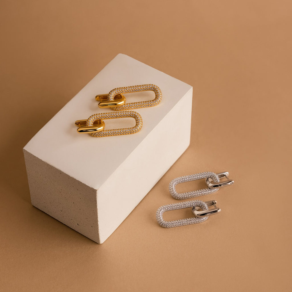 Two pairs of chain-link earrings—a pair of gold hoops and a pair of Pave Link Hoops—are displayed on and beside a white block against a beige background.
