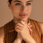 A freckled woman clasps her hands near her face, wearing a brown textured top and the Dainty Mama Ring against a beige background—a perfect minimalist ring and thoughtful gift for mom.