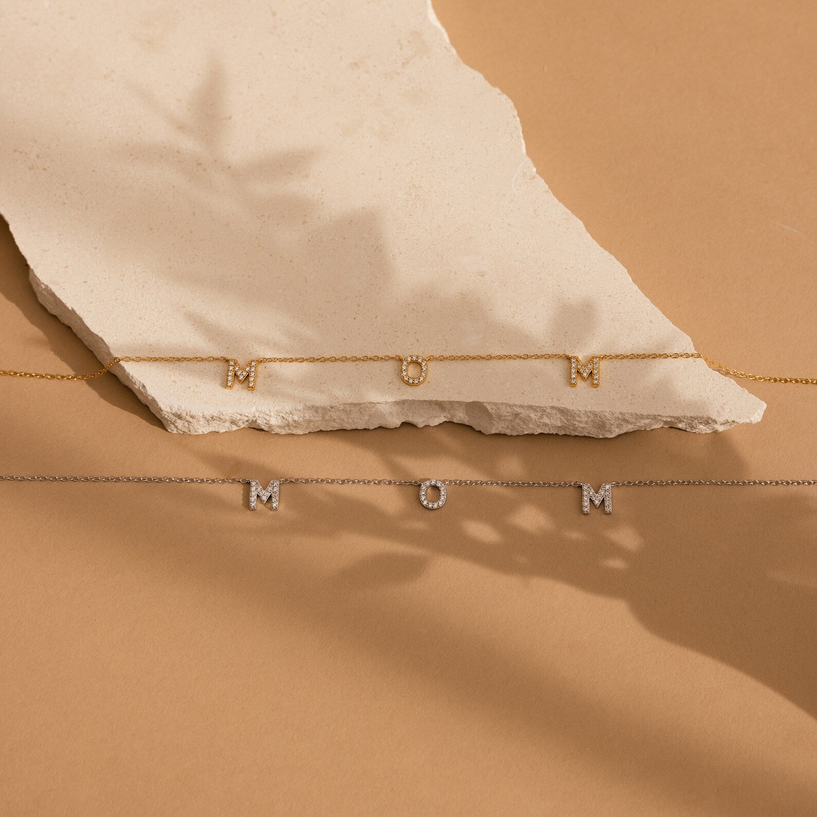 Two Pave Mom Necklaces—one in gold, one in silver—spell out "MOM" on a beige stone background. Each necklace is an elegant, meaningful gift for the special mom in your life.