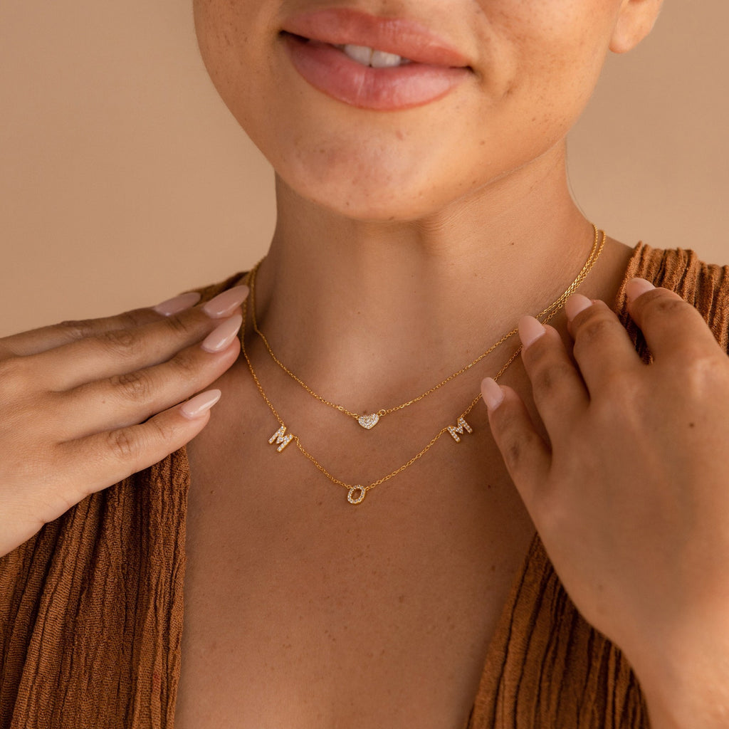 A woman wears the Pave Mom Necklace and a heart charm necklace, creating a thoughtful mom necklace duo that pairs beautifully with her brown top.
