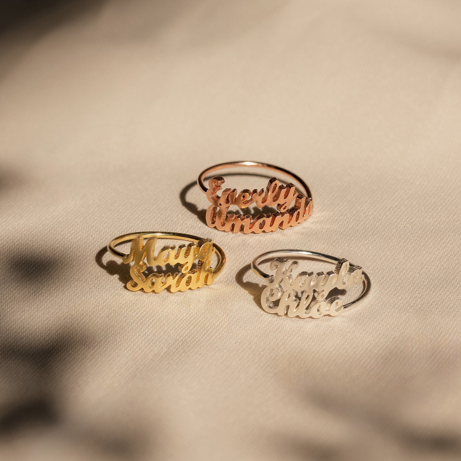 Three London Duo Name Rings in gold, rose gold, and silver are displayed on a beige fabric background.