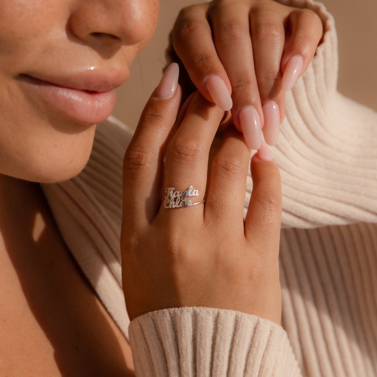 Close-up of a woman’s hand with long nails wearing the London Duo Name Ring personalized with “Tayla Chloe,” paired with a ribbed sweater.
