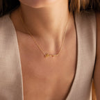 A woman in a beige sleeveless top wears the Tiny Venice Name Necklace, featuring a gold nameplate charm.