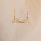 The Elegant Pave Name Necklace features "Brooke" in cursive, encrusted with sparkling gems and suspended from a chic twist chain for a personalized touch.