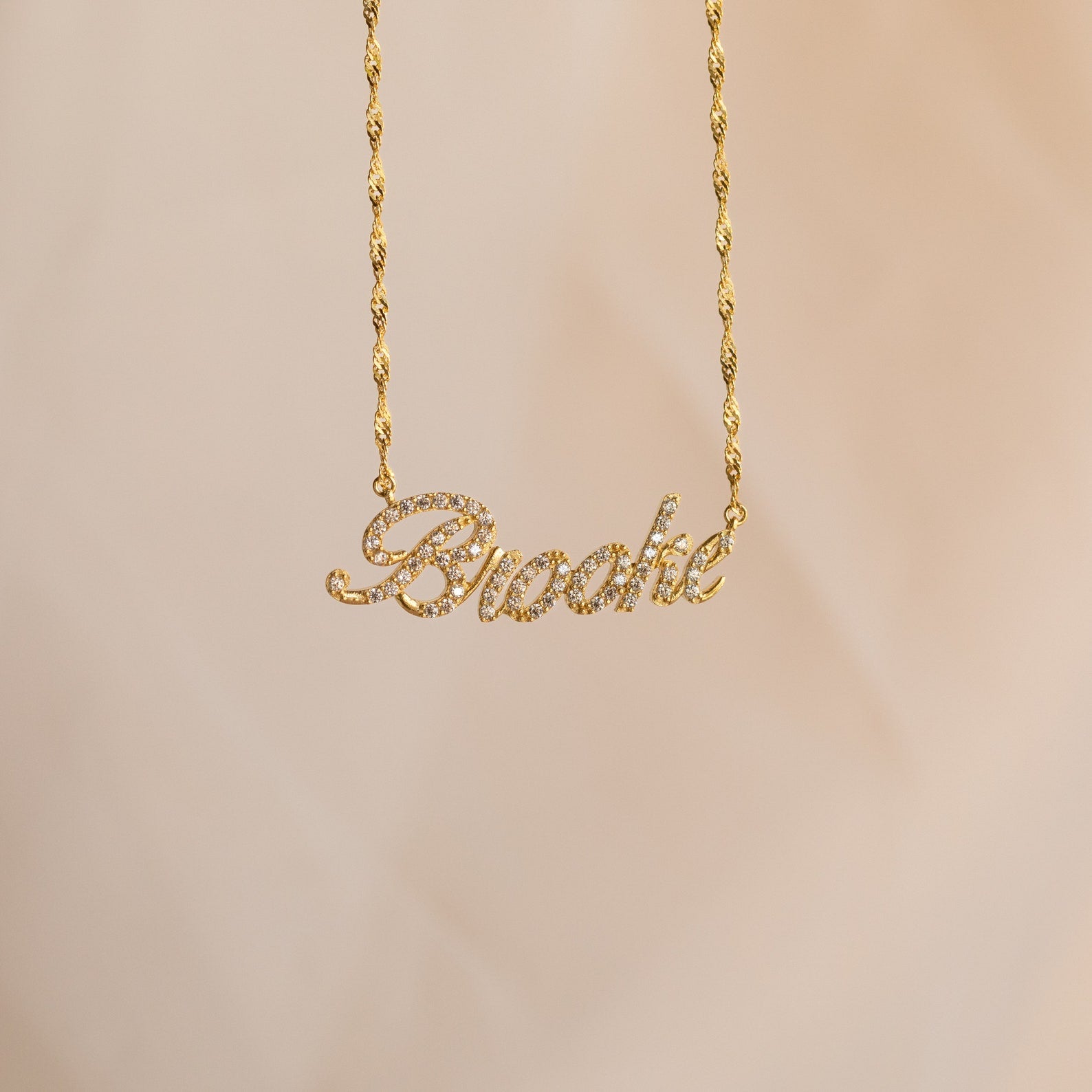The Elegant Pave Name Necklace features "Brooke" in cursive, encrusted with sparkling gems and suspended from a chic twist chain for a personalized touch.