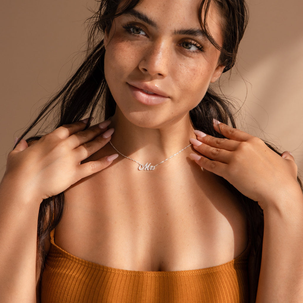 A woman with long dark hair in an orange top smiles as she touches her collarbone, wearing an Elegant Pave Name Necklace.