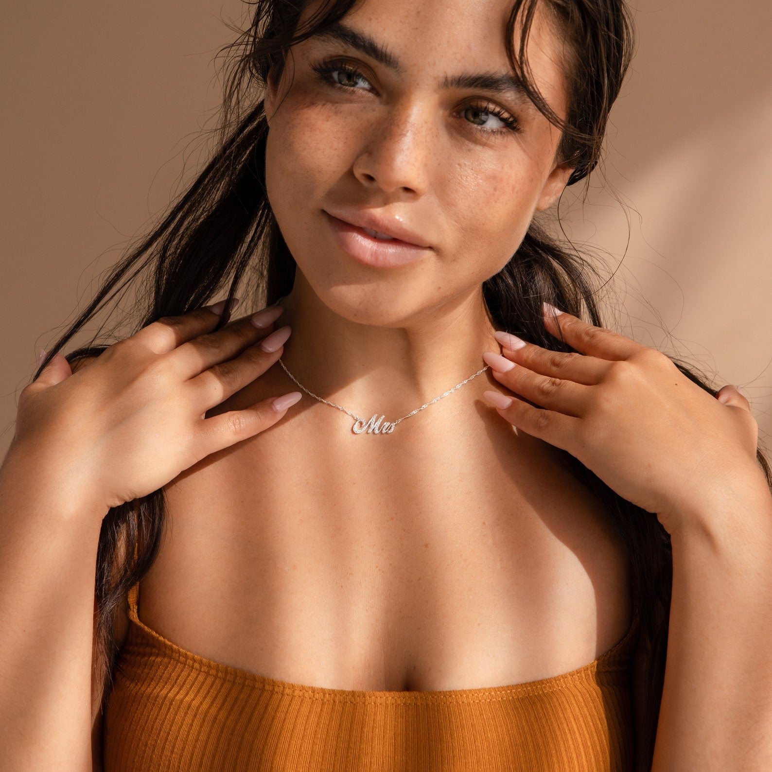 A woman with long dark hair in an orange top smiles as she touches her collarbone, wearing an Elegant Pave Name Necklace.
