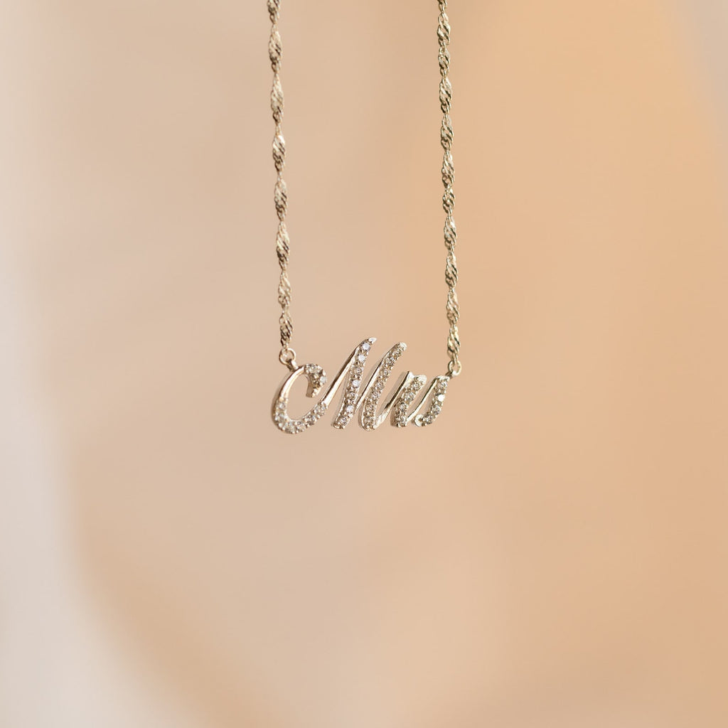 The Elegant Pave Name Necklace features a cursive "Mrs" pendant on a delicate twist chain, showcased against a soft beige background—a charming personalized accessory.