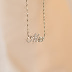 The Elegant Pave Name Necklace features a cursive "Mrs" pendant on a delicate twist chain, showcased against a soft beige background—a charming personalized accessory.