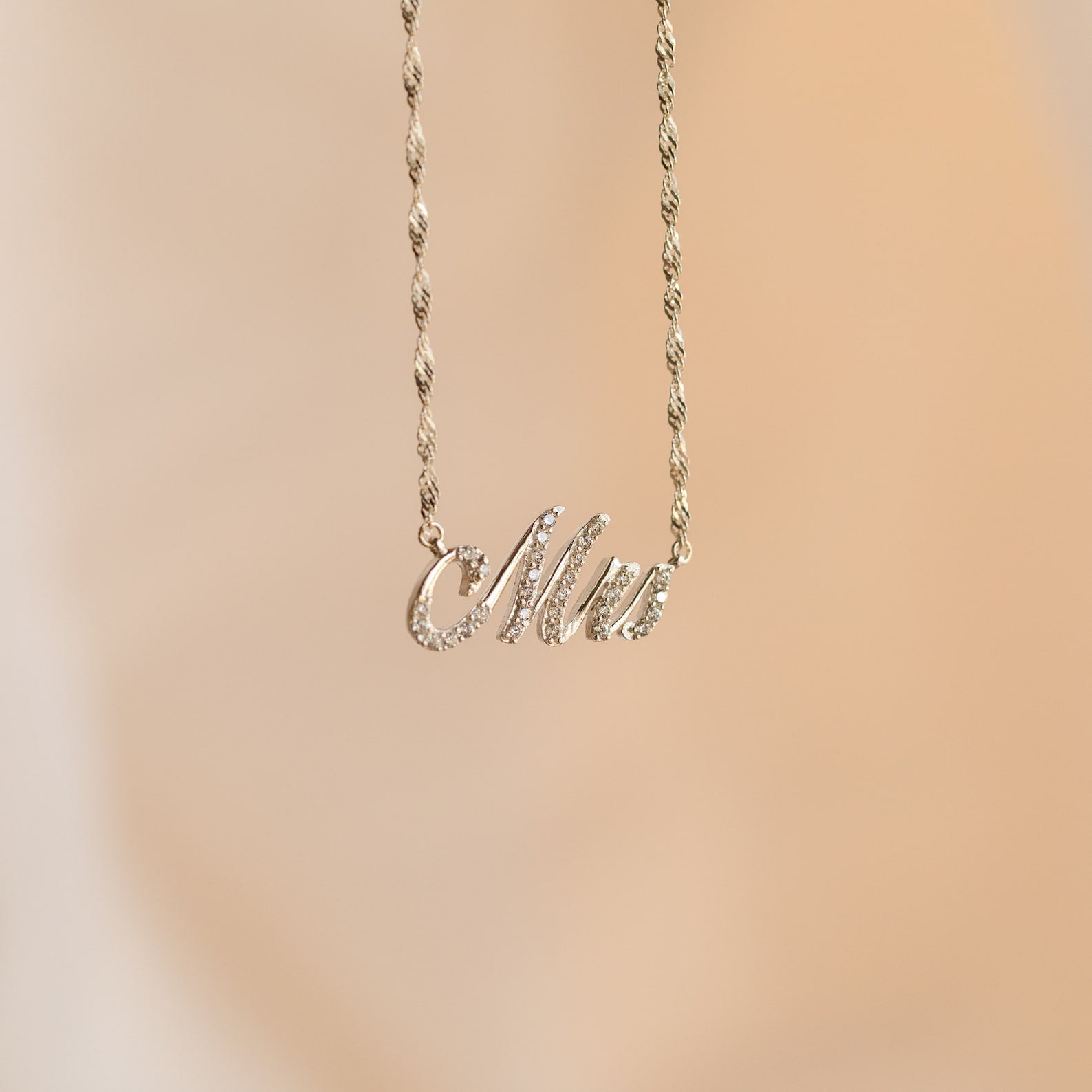 The Elegant Pave Name Necklace features a cursive "Mrs" pendant on a delicate twist chain, showcased against a soft beige background—a charming personalized accessory.
