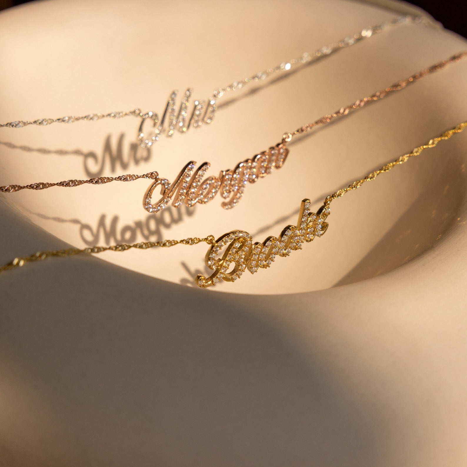 The Elegant Pave Name Necklace glimmers in sunlight on a beige curved surface, elegantly displayed alongside personalized nameplate necklaces—Mimi, Morgan, and Brooke.