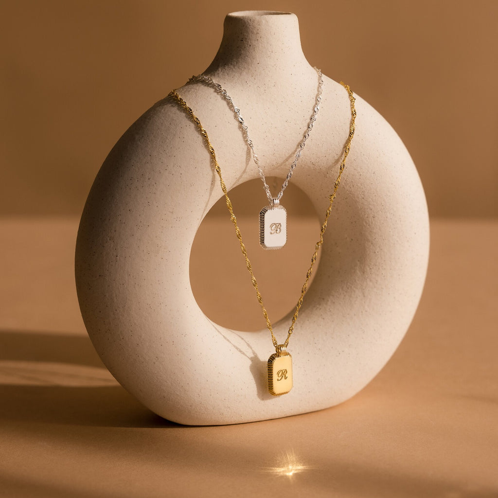Two Traditional Ribbed Initial Necklaces, one in silver and one in gold, with rectangular pendants, are elegantly displayed on a round white ceramic vase.