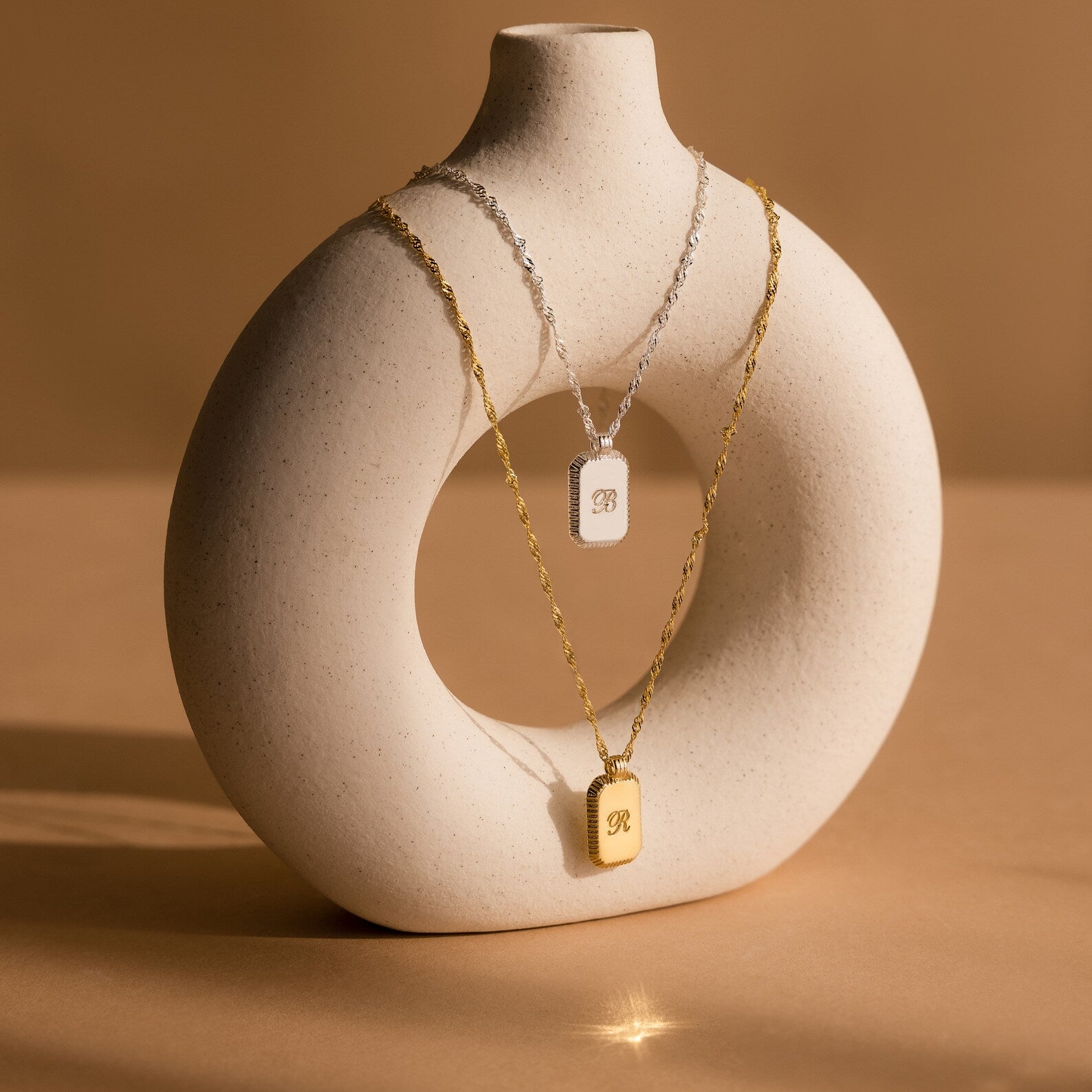 Two Traditional Ribbed Initial Necklaces, one in silver and one in gold, with rectangular pendants, are elegantly displayed on a round white ceramic vase.