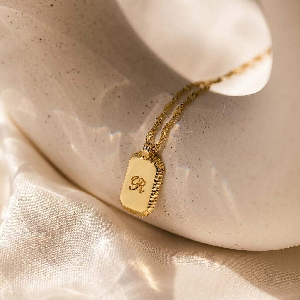 A gold Traditional Ribbed Initial Necklace featuring a rectangular pendant with the letter R rests against a smooth, pale surface.