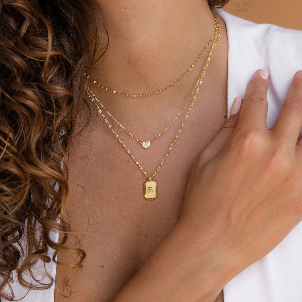 A woman wears the Traditional Ribbed Initial Necklace, a gold charm necklace featuring three layered chains with an engraved initial and a rectangle pendant, each pendant resting gently on her chest.