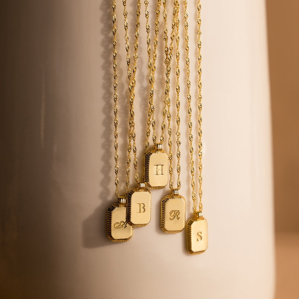 Traditional Ribbed Initial Necklaces rest on a light surface, each rectangle pendant showcasing a unique letter for a personalized charm look.