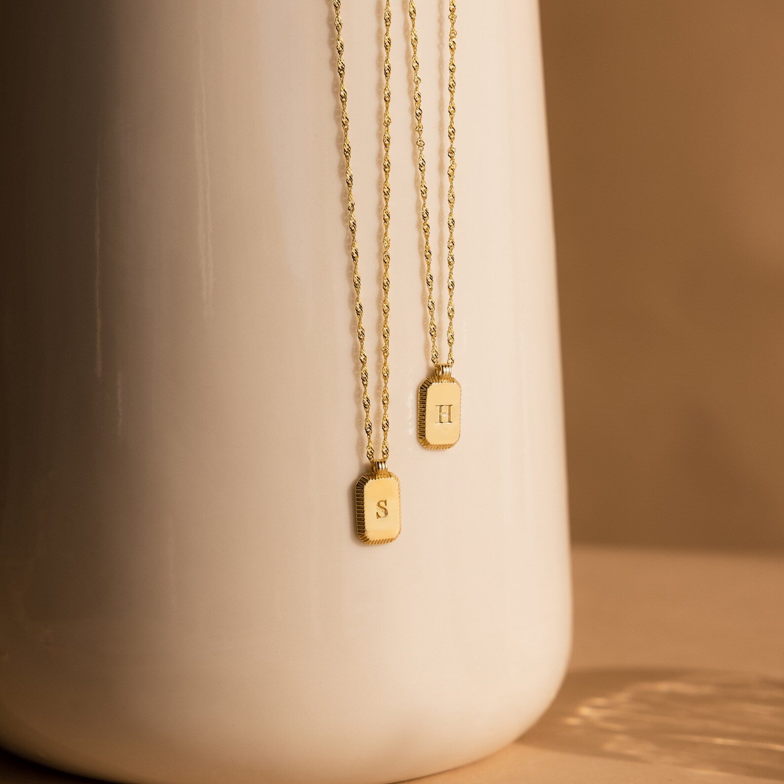 Two Newsletter Ribbed Initial Necklaces, one featuring an S and the other an H, hang gracefully on a white vase.