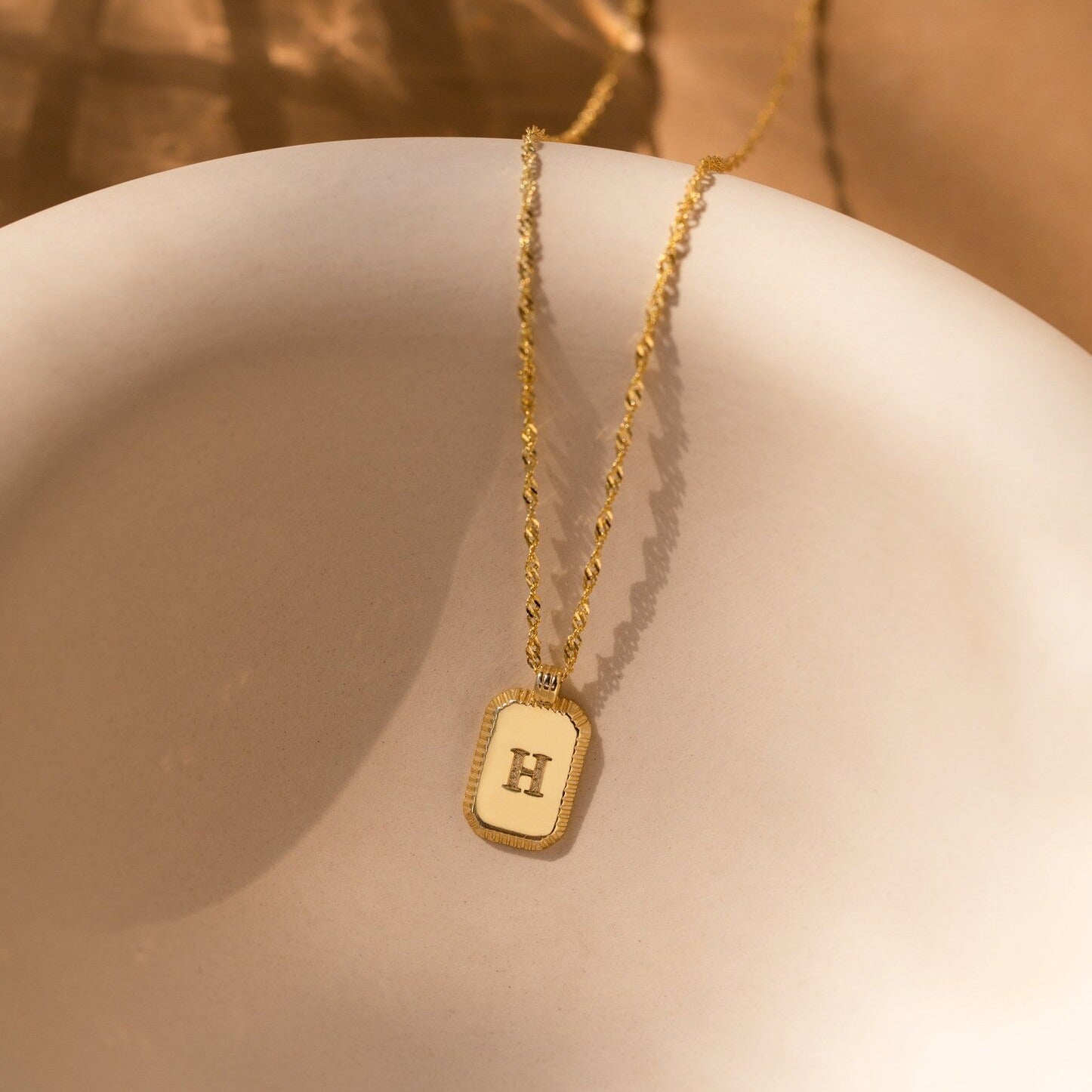 The Newsletter Ribbed Initial Necklace, engraved with the letter H, lies on a smooth white surface—a timeless charm perfect for showcasing personal style.