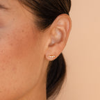 Close-up of a woman’s ear wearing Tiny Diamond Climbers—elegant small gold earrings with three white gemstones—perfect for anniversaries or as a thoughtful gift.