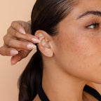 A woman with freckles touches her ear, highlighting the elegant Tiny Diamond Climbers—curved earrings adorned with small diamonds, making them a perfect anniversary gift.