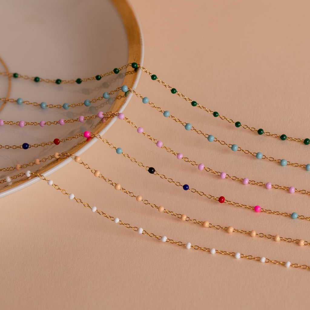 The Peach Station Necklace and five other delicate gold chain necklaces with colorful beads are displayed on a peach background and white dish, perfect for layering to achieve a beach-inspired look.