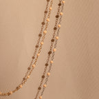 The Peach Station Necklace, featuring gold and black beads, drapes elegantly on a beige textured fabric—perfect for adding a stylish layer and elevating your beach-inspired look.