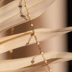The Peach Station Necklace features gold chain and small beige beads, shown draped over cream dried palm leaves in soft sunlight—perfect for a beachy look or layering.