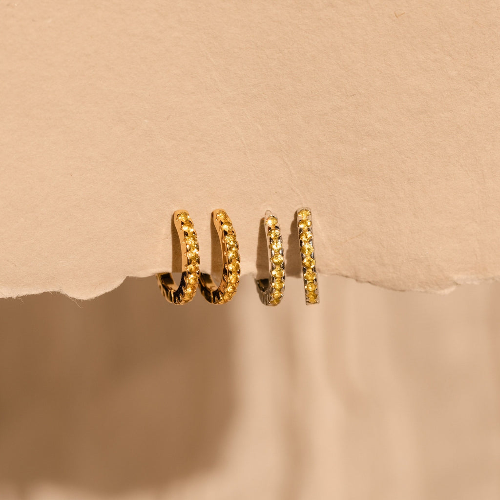 Three Citrine Pave Huggies hang elegantly on a torn piece of beige paper, their textured gold surfaces sparkling as they catch the light.