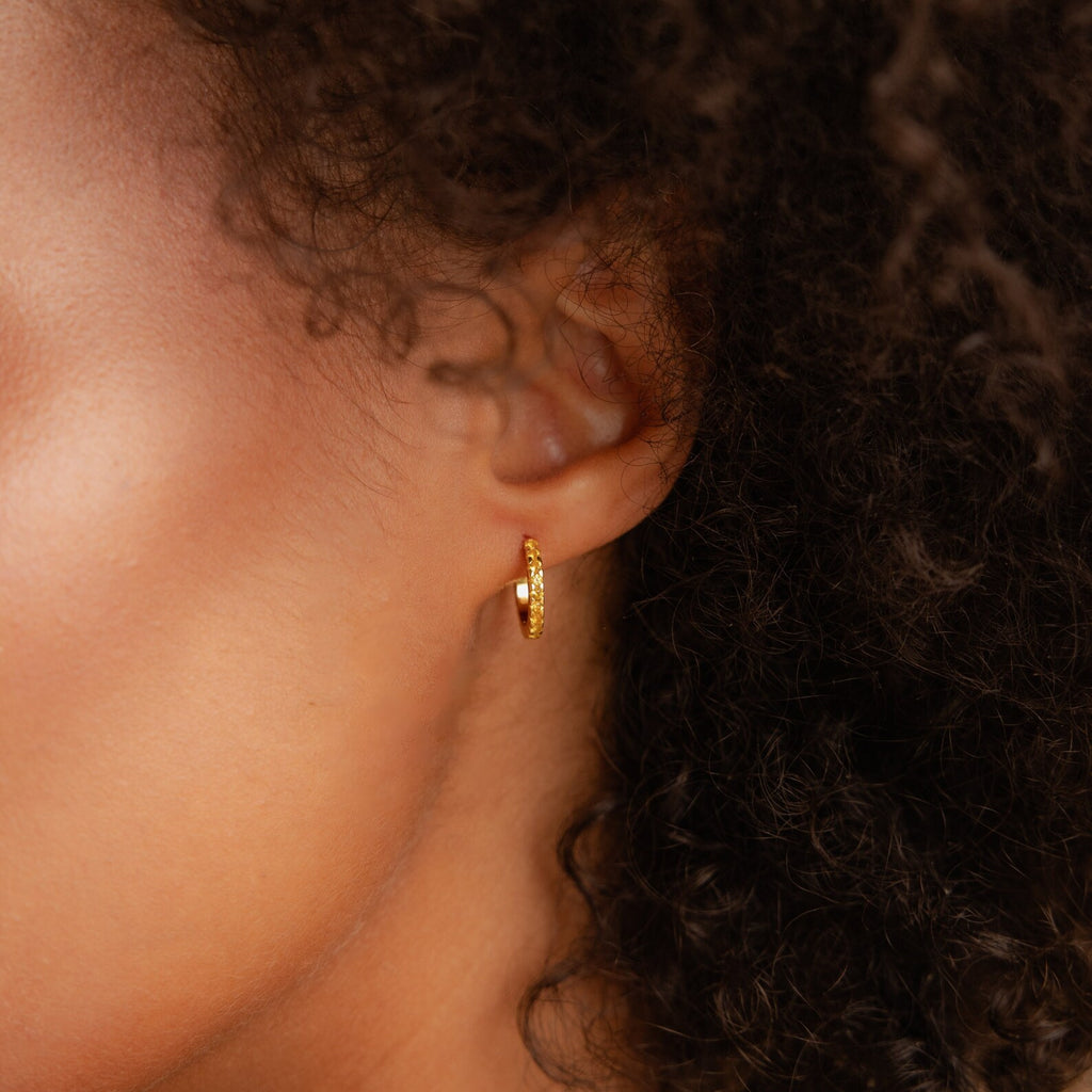 Close-up of a person’s ear adorned with Citrine Pave Huggies, showcasing small hoop earrings, curly hair, and smooth skin.