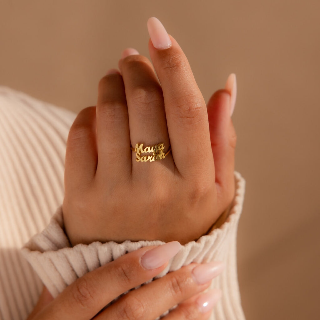 A manicured hand displays the London Duo Name Ring in gold, featuring personalized engraved names, set against a beige background.