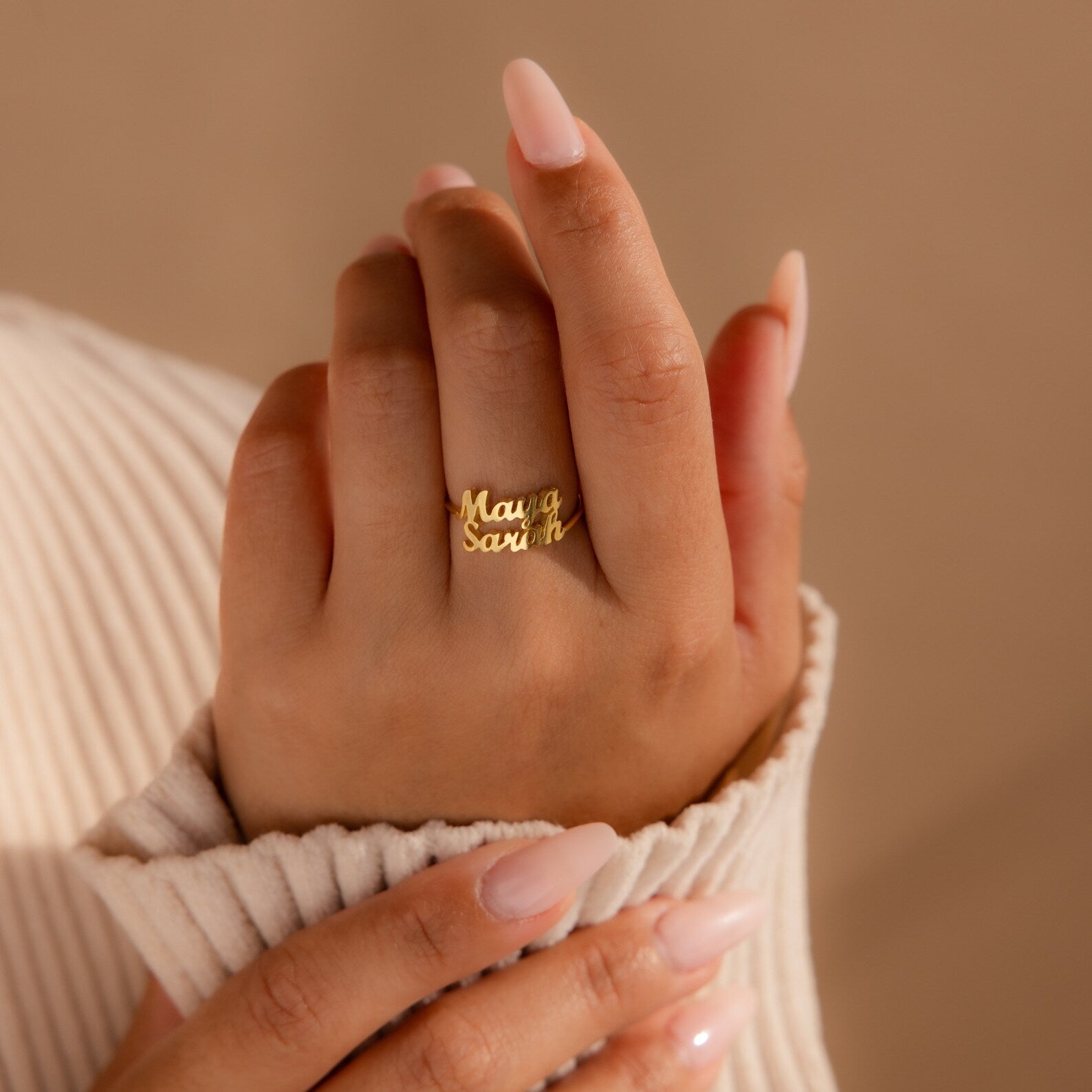 A manicured hand displays the London Duo Name Ring in gold, featuring personalized engraved names, set against a beige background.