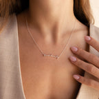 A woman in a beige top touches her collarbone while wearing the Tiny Venice Name Necklace with "kimberly" in cursive.