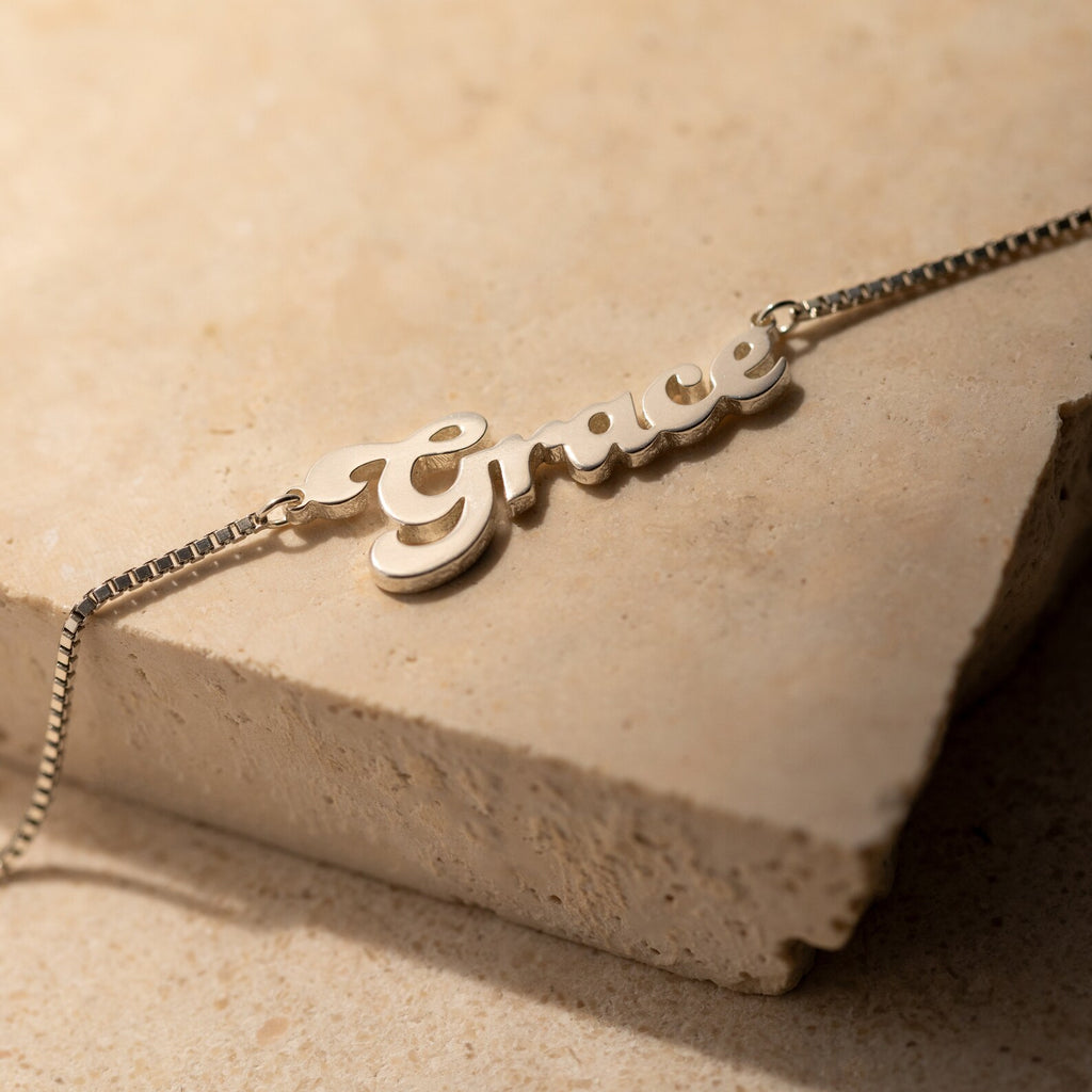 The Groovy Name Necklace in Box Chain, personalized with the name Grace in elegant cursive, is displayed beautifully on a beige stone surface.