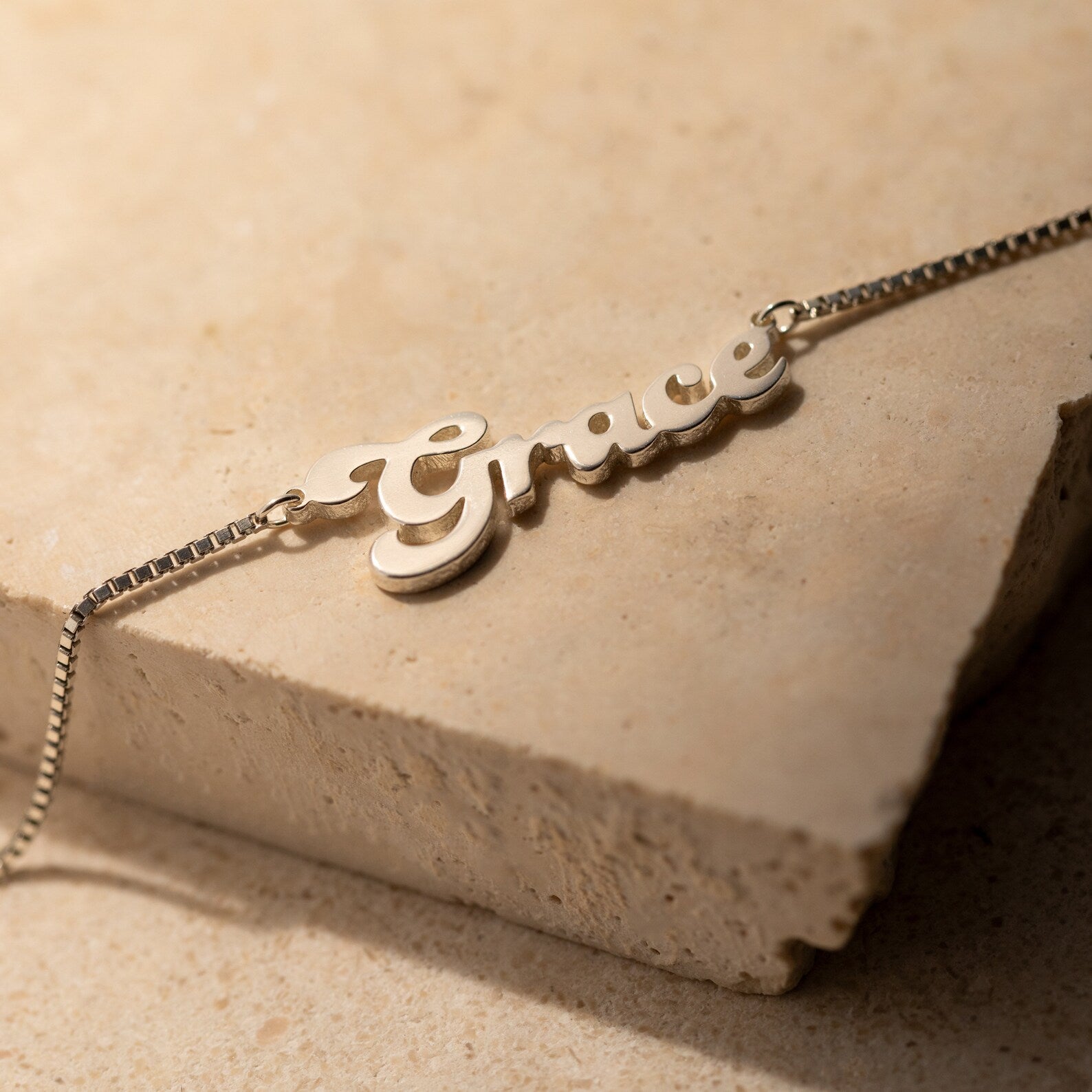 The Groovy Name Necklace in Box Chain, personalized with the name Grace in elegant cursive, is displayed beautifully on a beige stone surface.