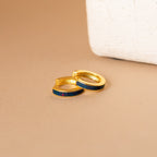 Opal Inlay Earrings with striking blue and red opal inlays set in gold hoops are displayed on a beige surface beside a white object. Part of our Opal Collection, they bring vibrant color to any outfit.