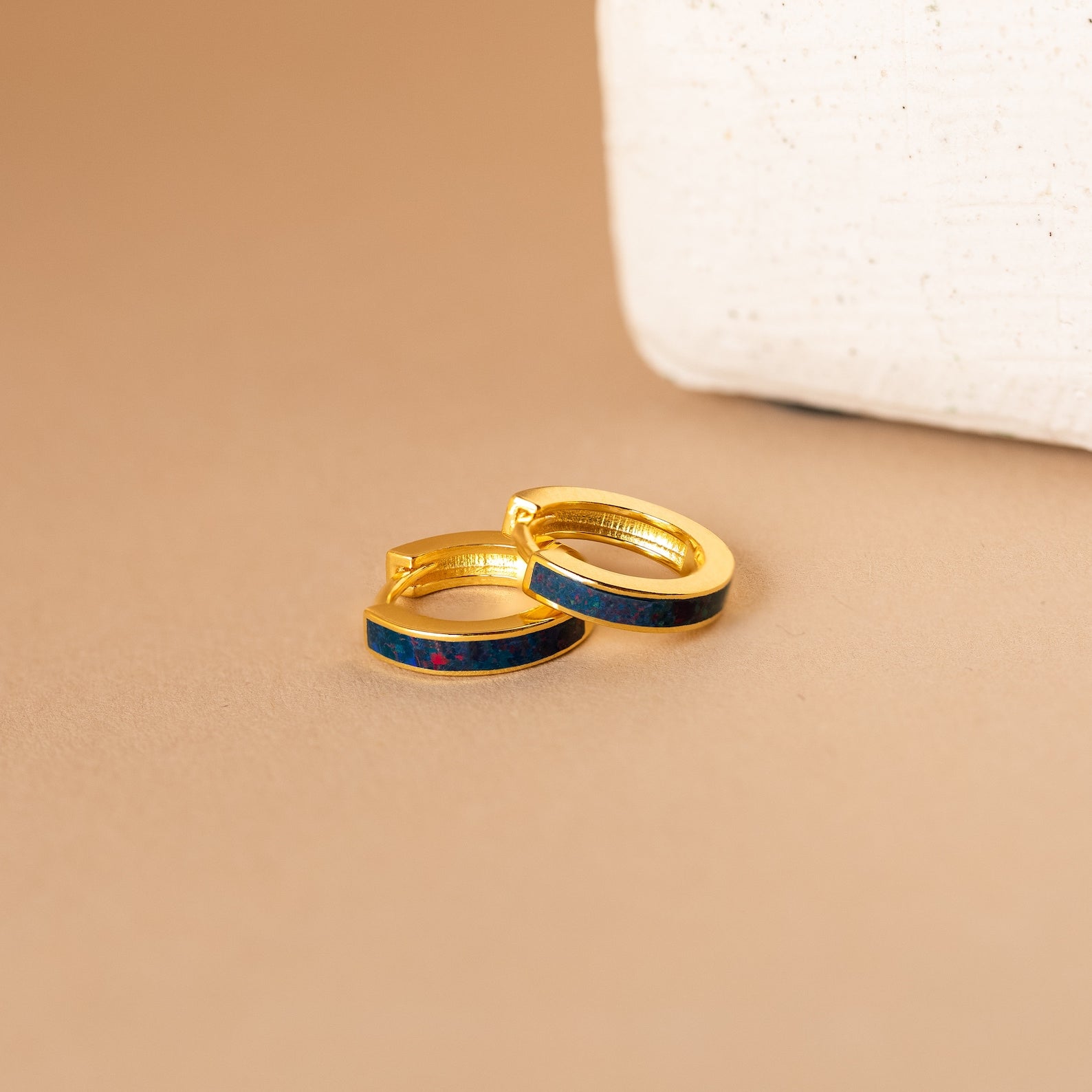 Opal Inlay Earrings with striking blue and red opal inlays set in gold hoops are displayed on a beige surface beside a white object. Part of our Opal Collection, they bring vibrant color to any outfit.
