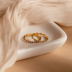 The Aquamarine Eternity Ring, a gold stacking ring adorned with aquamarine gemstones, rests on a white ceramic dish beside sheer beige fabric.