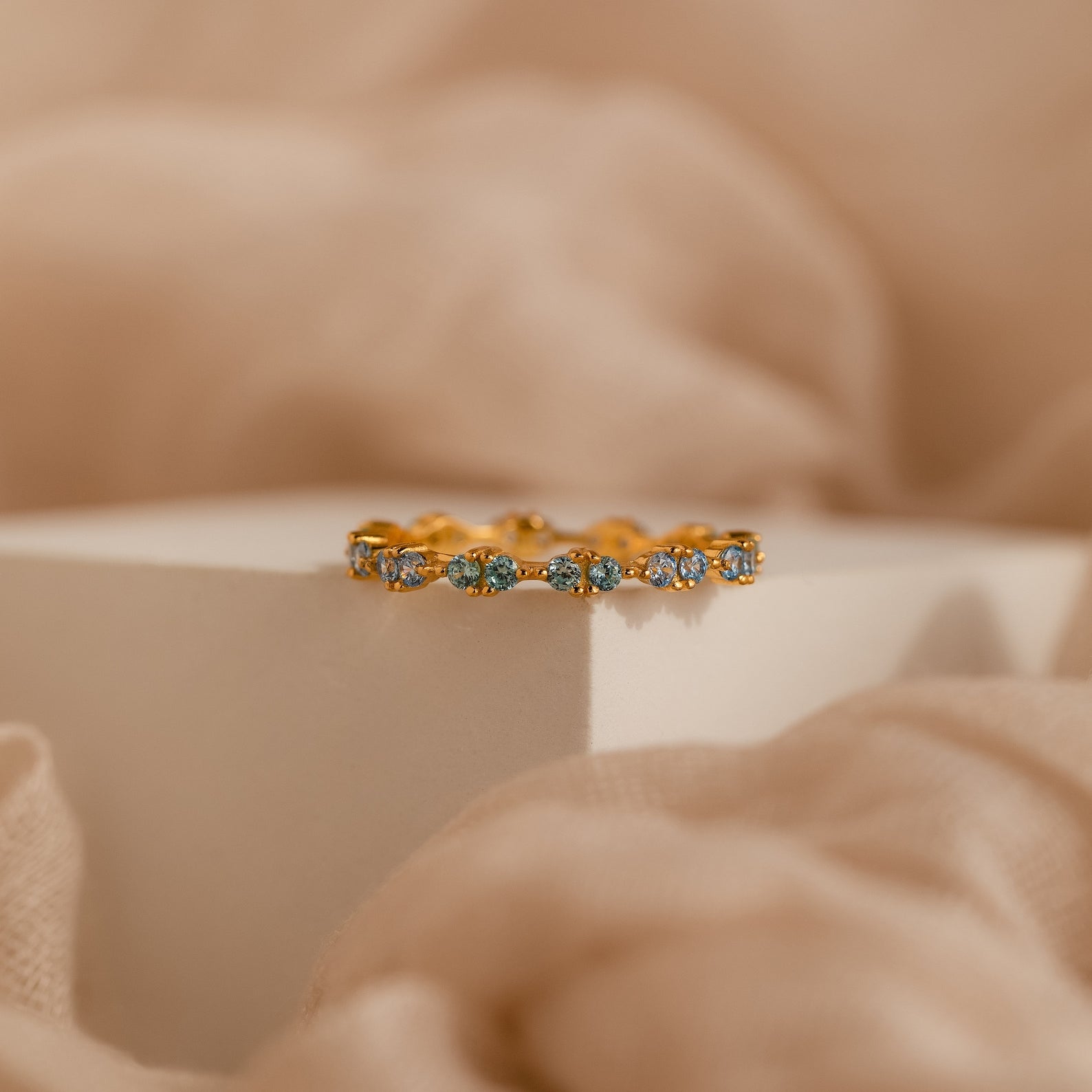 The Aquamarine Eternity Ring, featuring shimmering blue gemstones, is displayed on a white block and surrounded by soft beige fabric.