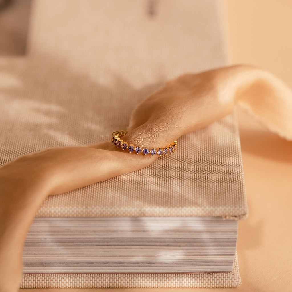 The Amethyst Eternity Ring pairs beautifully with a gold bracelet featuring purple amethyst gemstones, displayed on a beige book with soft tan fabric—ideal for an elegant, timeless style.