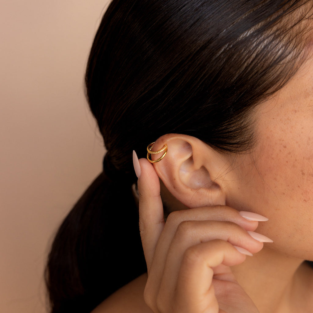 A woman with a low ponytail touches her manicured ear, adorned with multilayered earrings and the Lydia Duo Cuffs in gold.
